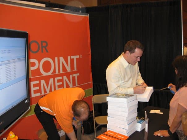 Errin O'Connor signing a stack of his Windows SharePoint Services 3.0 Inside Out (Microsoft Press) book at the Boston SharePoint Technology Conference, 2010 — orange POINT MANAGEMENT branded booth in background, attendee in white blouse waiting, live SharePoint dashboard on the adjacent monitor